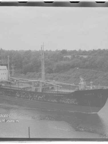 Schwarzweißaufnahme eines Frachtschiffs mit verwittertem Rumpf, das in einem Kanal an bewaldetem Ufer vorbeifährt; sichtbarer Filmrand und Wasserzeichen des Internationalen Maritimen Museums Hamburg unten links.