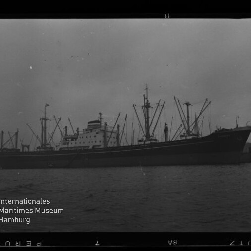 Schwarz-weiß-Foto eines großen Frachtschiffs mit zahlreichen Ladekränen und Masten am Kai unter trübem Himmel; Wasser im Vordergrund, Filmrand und Museumslogo des Internationalen Maritimen Museums Hamburg unten links erkennbar.