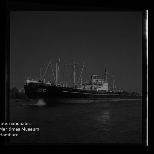 Großes Frachtschiff mit mehreren Masten und Ladekränen fährt auf ruhigem Wasser bei dunklem Himmel, mit sichtbarer Bugwelle, Filmrahmenrand und Wasserzeichen des Internationalen Maritimen Museums Hamburg unten links.