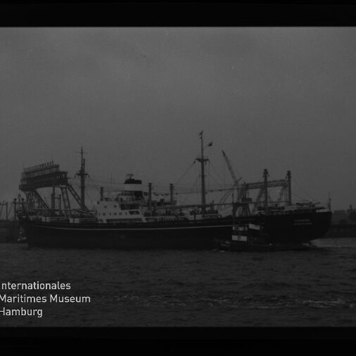 Schwarzweißaufnahme eines größeren Frachtschiffs auf dem Wasser mit einem kleinen Schlepper entlang der Seite, im Hintergrund Hafenkräne und Industrieanlagen; Museumssignet ‚Internationales Maritimes Museum Hamburg‘ unten links.