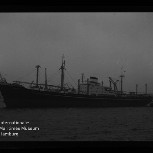 Schwarzweißaufnahme eines langen Frachtschiffs mit mehreren Ladekränen und Masten, im Hafen vor dunklem Wasser liegend, mit sichtbarem Filmrand und Museumssignet in der linken unteren Ecke.