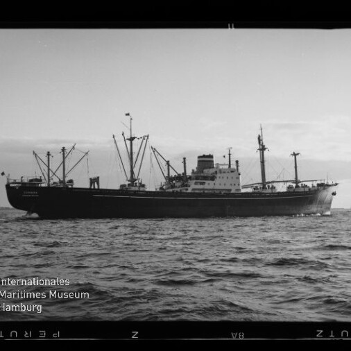 Schwarzweißaufnahme eines Frachtschiffs mit mehreren Ladekränen und mittig gelegenem Aufbau auf ruhiger See vor flachem Horizont; Filmrand und Wasserzeichen des Internationalen Maritimen Museums Hamburg in der unteren linken Ecke sichtbar.