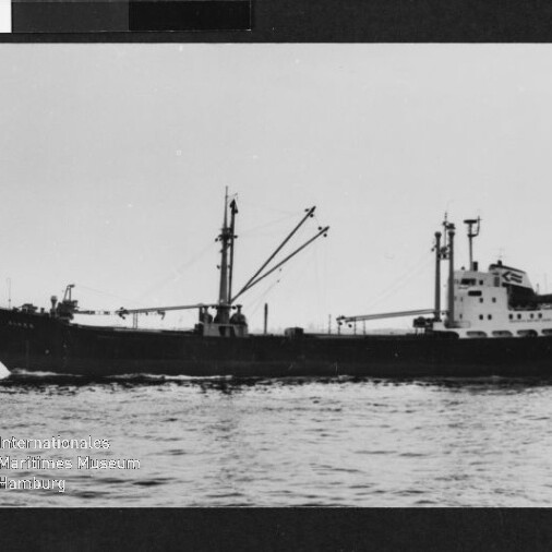 Schwarzweiß-Seitenansicht eines Frachtschiffs mit zwei Masten und Ladebäumen sowie achterlicher Aufbauten, das ruhig auf offener See fährt; unten links Wasserzeichen des Internationalen Maritimen Museums Hamburg.