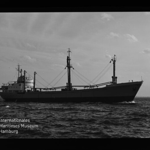 Schwarz-weiße Aufnahme eines Frachtschiffs in Seitenansicht auf der See mit zwei Masten und Ladegeschirren, dunklem Filmrand und dem Wasserzeichen des Internationalen Maritimen Museums Hamburg.
