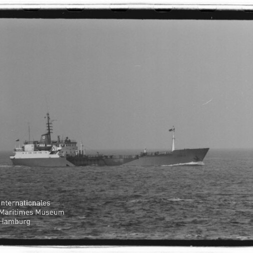 Schwarz-weiß-Foto eines langen, niedrig bauenden Frachtschiffs mit Aufbauten mittschiffs, Masten und Flagge am Bug, das auf ruhiger See fährt; unten links das Logo des Internationalen Maritimen Museums Hamburg.