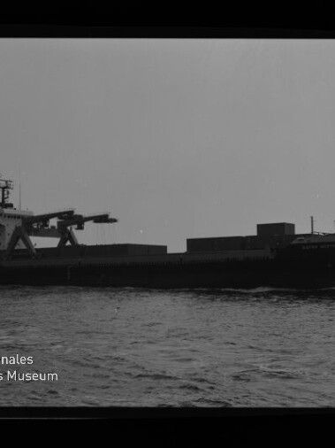 Schwarz-weiß-Seitenansicht eines Frachtschiffs mit markantem Ladekran und aufgestapelten Ladeeinheiten, das ruhig über das Meer vor hellem Himmel fährt; unten links das Logo und der Schriftzug des Internationalen Maritimen Museums Hamburg.