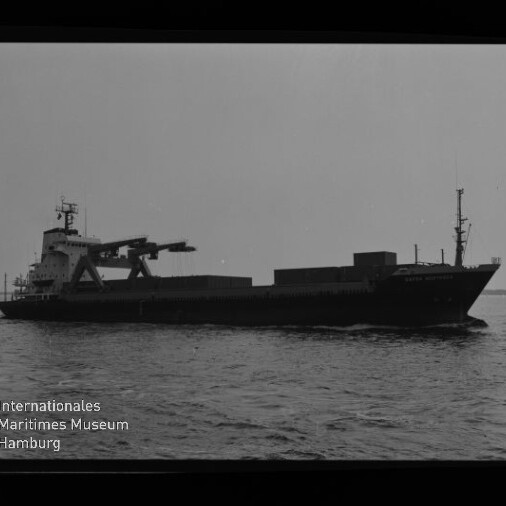 Schwarz-weiß-Seitenansicht eines Frachtschiffs mit markantem Ladekran und aufgestapelten Ladeeinheiten, das ruhig über das Meer vor hellem Himmel fährt; unten links das Logo und der Schriftzug des Internationalen Maritimen Museums Hamburg.