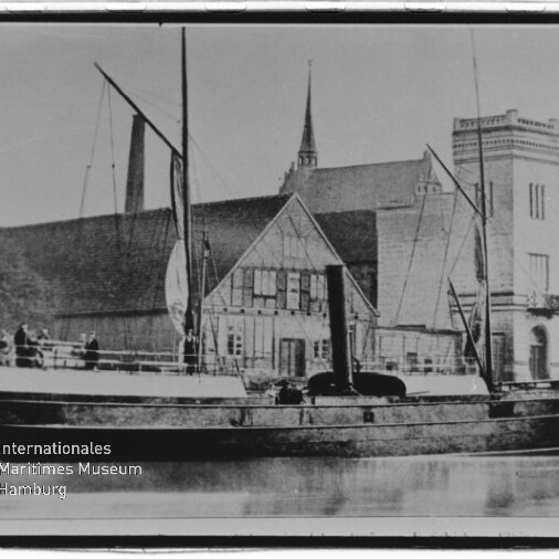 Schwarzweißaufnahme eines historischen Dampfschiffs mit Schornstein, Masten und aufgeholten Segeln, das am Kai vor einem Fachwerkgebäude und einem großen Backsteinhaus liegt, mit Spiegelung im ruhigen Wasser und einigen Personen auf dem Deck.