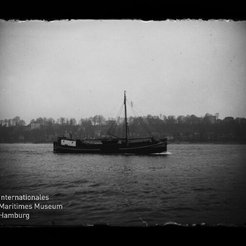 Schwarzweiß-Foto eines einmastigen Frachtschiffs, das ruhig auf dem Wasser vor einem baumbestandenen, nebligen Ufer mit Gebäuden fährt; unten links das Logo des Internationalen Maritimen Museums Hamburg.