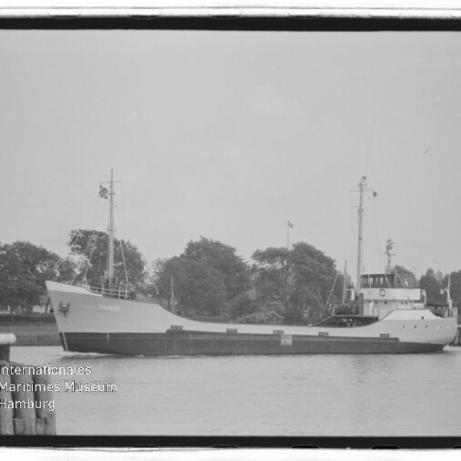 Schwarzweißaufnahme eines Frachtschiffs in Seitenansicht mit niedrigem Laderaum, zwei Masten und achterlichem Steuerhaus, vor einem baumbestandenen Ufer auf ruhigem Wasser.