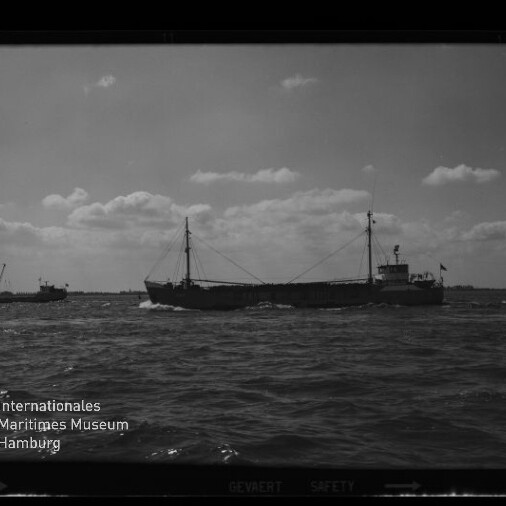 Schwarzweißaufnahme eines Frachtschiffs mit zwei Masten und achterlichem Aufbau, das auf leicht bewegter See fährt; links ein kleines Begleitschiff, rechts ein Segelboot am Horizont und unten links das Logo des Internationalen Maritimen Museums Hamburg.