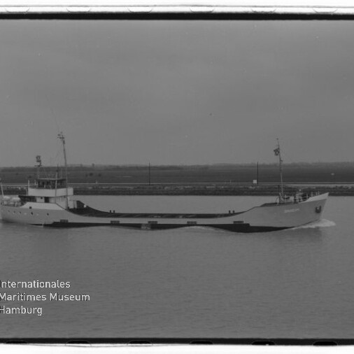Schwarzweiß-Foto eines flachen Frachtschiffs, das in ruhigem Gewässer an einem flachen Ufer vorbeifährt; links im Vordergrund ein kleines Segelboot und in der unteren linken Ecke ein Wasserzeichen des Internationalen Maritimen Museums Hamburg.