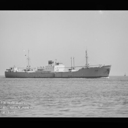 Schwarzweiß-Seitenansicht eines Frachtschiffs auf ruhigem Meer vor flachem Horizont, mit sichtbarem Filmrand, Museumswasserzeichen unten links und einem kleinen Segelboot in der Ferne rechts.
