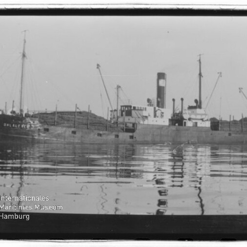 Schwarzweiß-Foto eines alten Frachtschiffs mit markantem Schornstein, mehreren Masten und Ladekränen, ruhig im Wasser liegend mit klarer Spiegelung und dem Wasserzeichen ‚Internationales Maritimes Museum Hamburg‘ links unten.