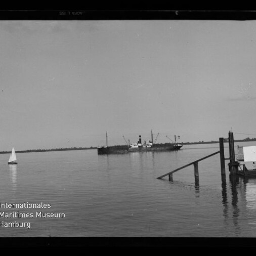 Ruhige Wasserfläche mit einem Dampfschiff in Bildmitte, einem kleinen Segelboot links und einem Pier mit einigen Personen rechts, flacher Horizont im Hintergrund.