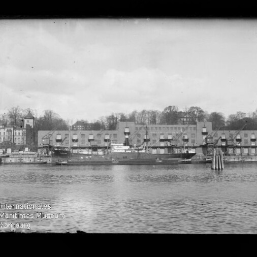 Blick über das Wasser auf einen Hafen mit mehreren angelegten Frachtschiffen vor einer langen Reihe alter Lagerhäuser und Gebäuden, im Hintergrund ein baumbewachsener Hang.