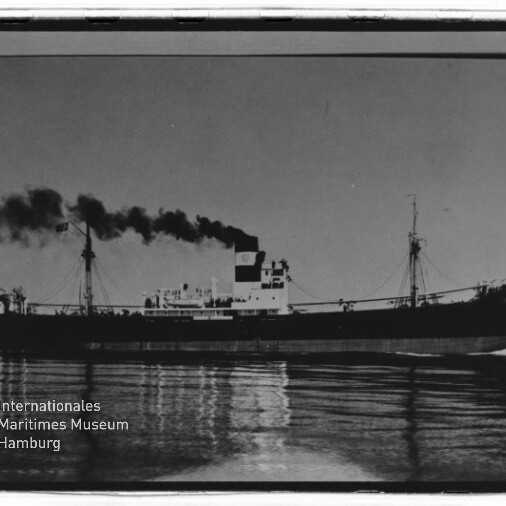 Schwarz-weiße Seitenaufnahme eines Frachtschiffs/Dampfers auf ruhigem Wasser mit hohem Schornstein, aus dem dunkler Rauch aufsteigt, und deutlicher Spiegelung im Wasser; unten links Stempel des Internationalen Maritimen Museums Hamburg.
