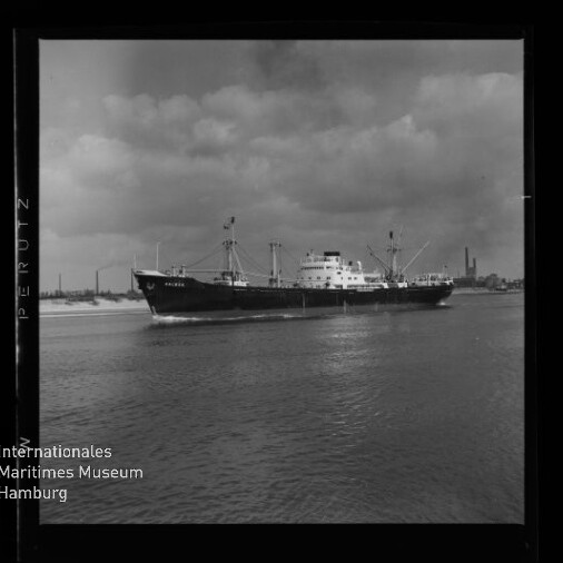 Schwarzweiß-Foto eines Frachtschiffs, das nahe einer Industriehafenpromenade über ruhiges Wasser fährt; wolkiger Himmel, deutlicher schwarzer Rahmen und Museumssiegel unten links.