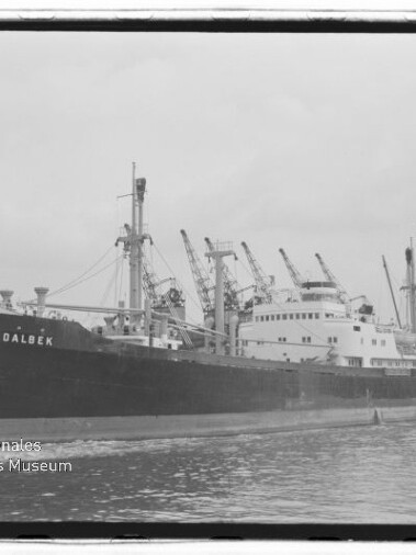 Schwarzweiß-Foto eines Frachtschiffs mit dem gut lesbaren Namen DALBEK am Bug, das am Hafen liegt, mit mehreren Ladebäumen und Hafenkränen im Hintergrund, ruhigem Wasser im Vordergrund und Museumslogo unten links.