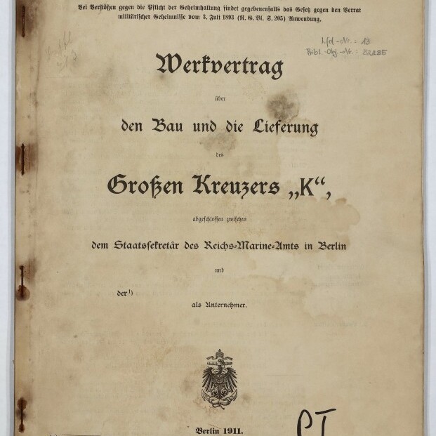 Deckblatt eines historischen Vertrags mit dem großen Titel „Werkvertrag über den Bau und die Lieferung des Großen Kreuzers ‚K‘“, oben handschriftlich in Rot „Vorentwurf“, gedruckt „Berlin 1911“ und ein preußischer Adler als Wappen, seitliche Rost- und Wasserflecken am Einband sowie Museumslogo unten links und handschriftliche Notiz „PT 13“ unten rechts.