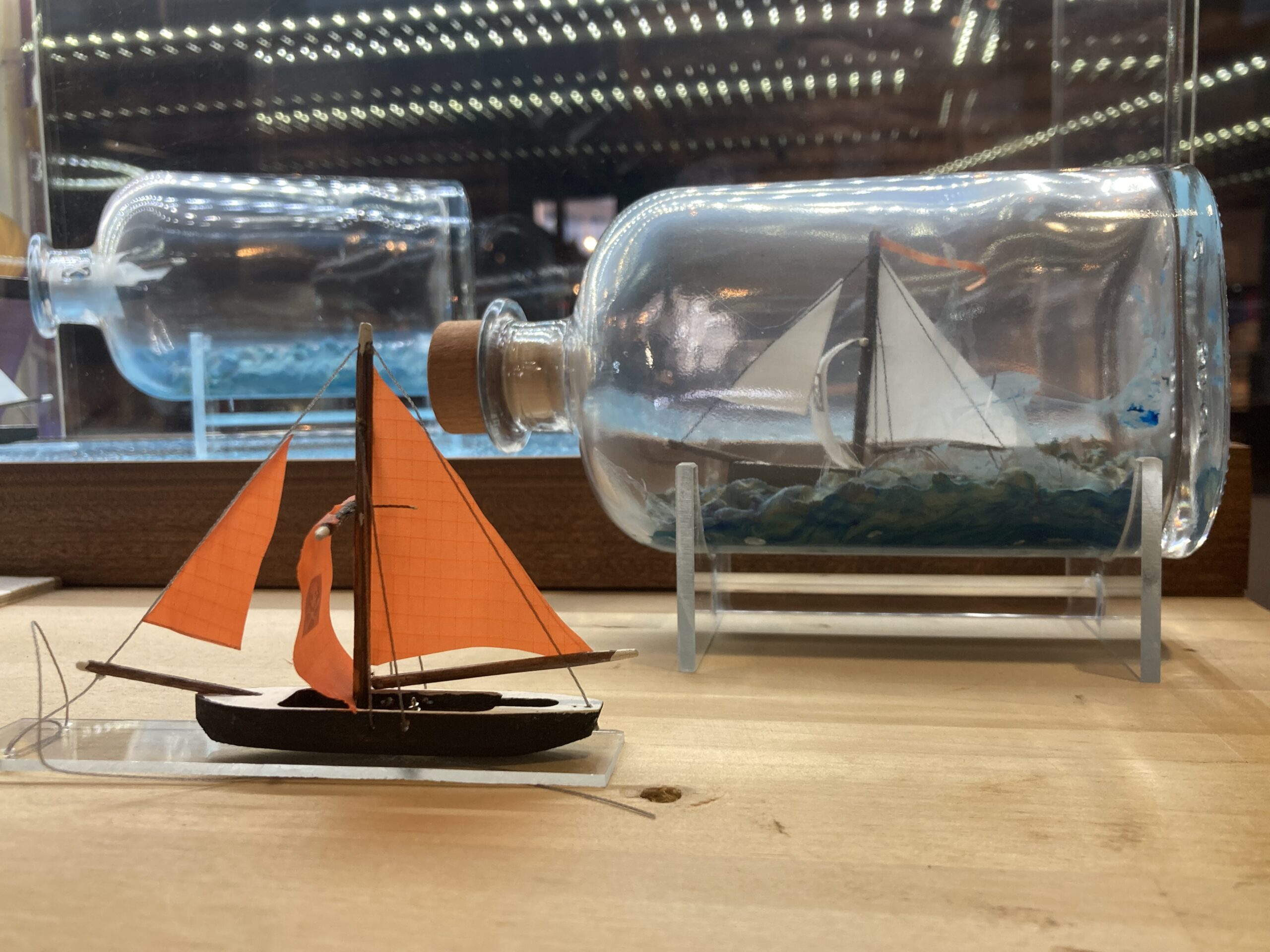Modellsegelboot mit leuchtend orangefarbenen Segeln im Vordergrund und ein weißes Segelmodell in einer Flasche auf einem Holzuntergrund.