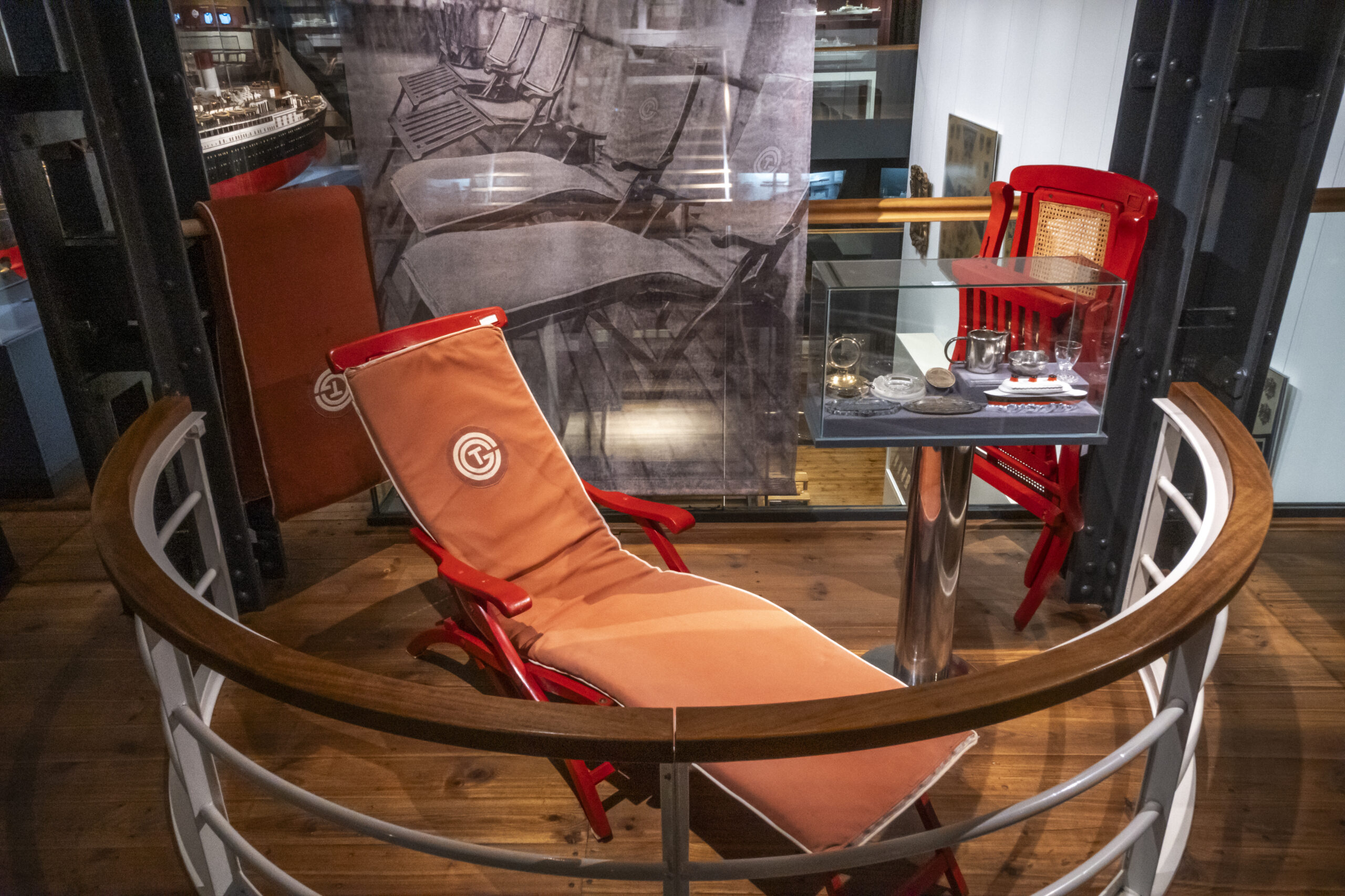 Roter Liegestuhl mit orangefarbenem Polster in einer Museumspräsentation, daneben eine Glasvitrine mit kleinem Tee-Service.