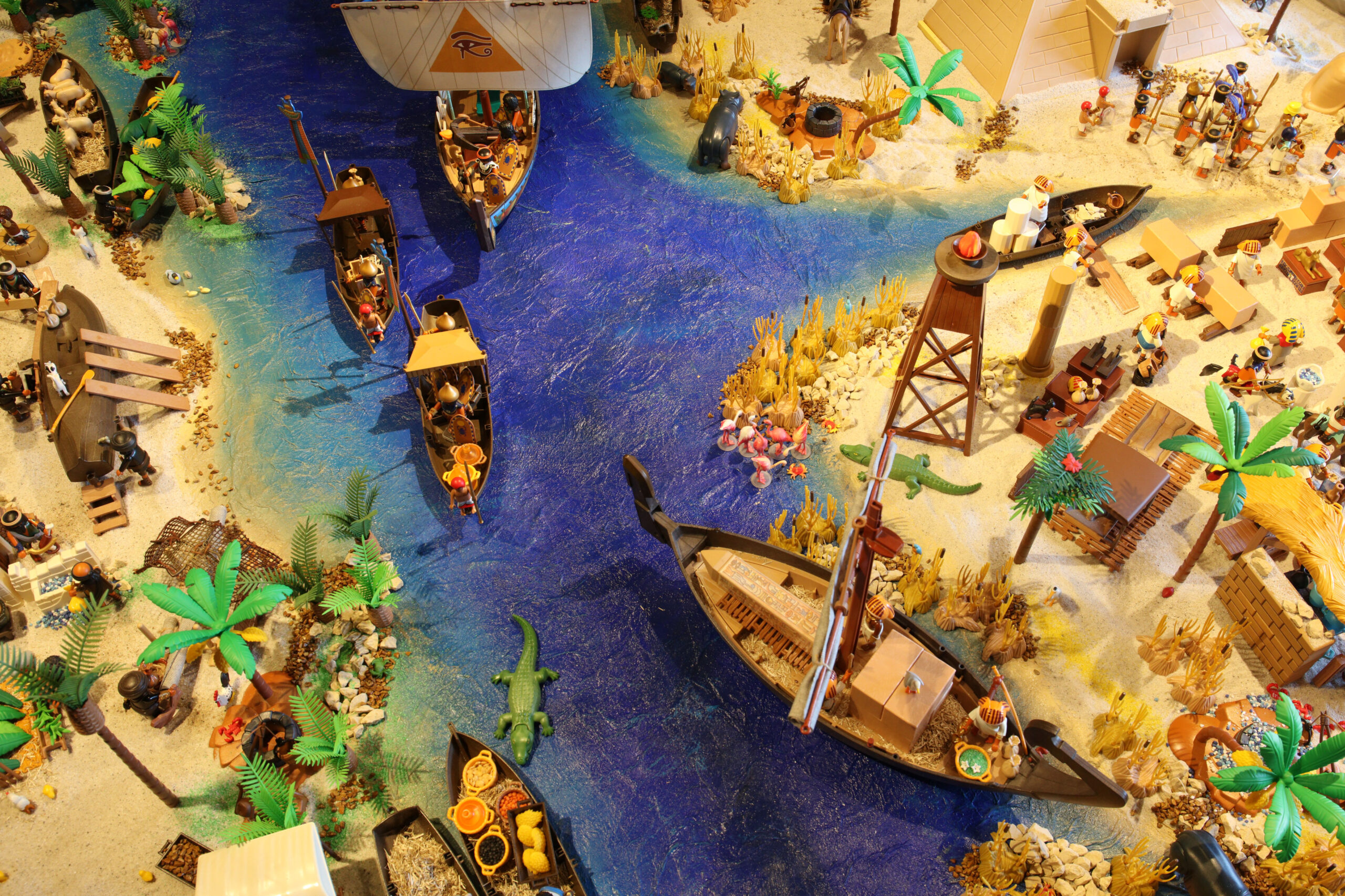 Spielzeug-Diorama einer Flussmündung mit blauen Gewässern, mehreren Booten, Palmen, Figuren und einer großen Ladung auf einem Segelschiff.