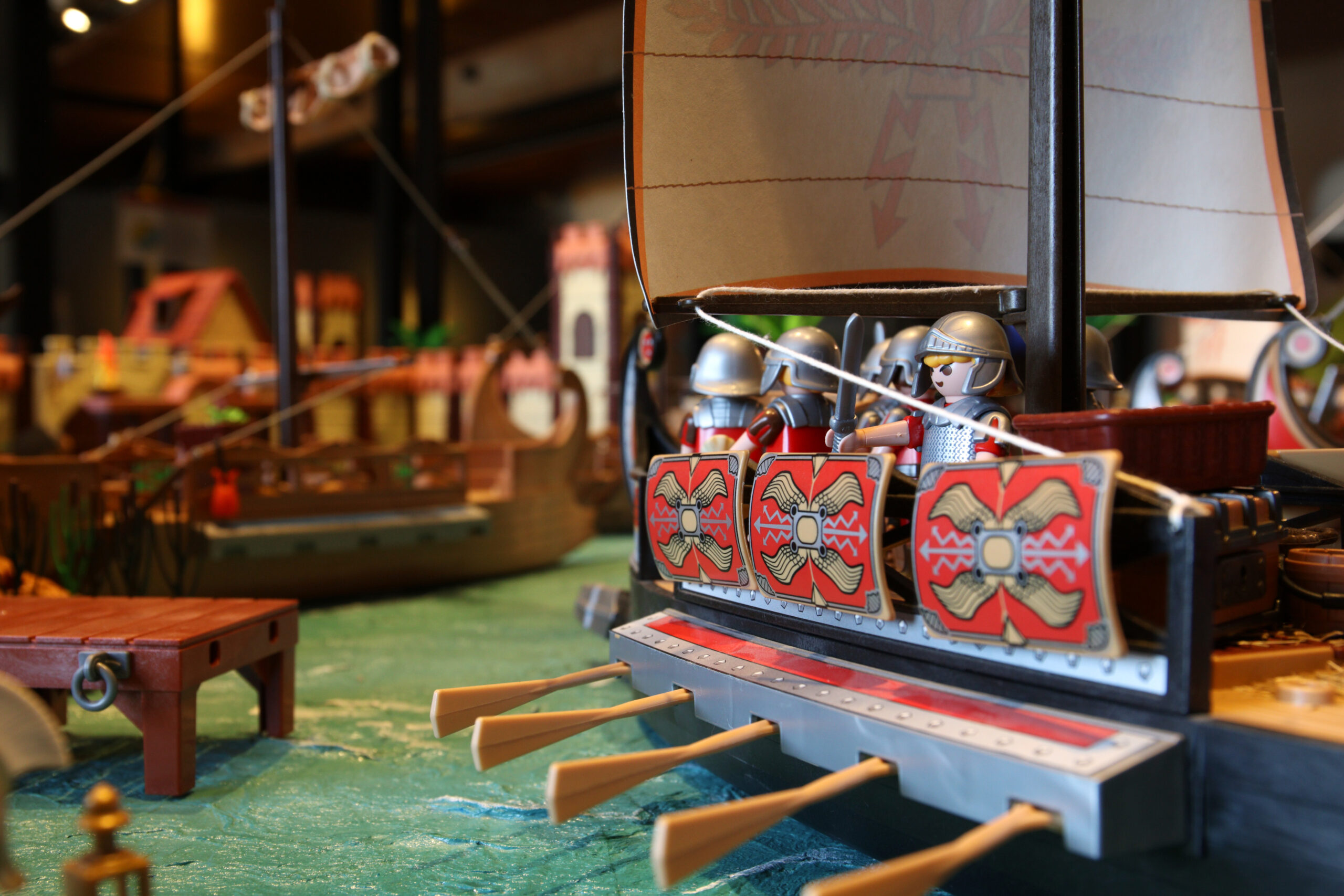 Spielzeug-Ruderboot mit bemalten Schilden, Playmobil-Rittern in Helmen und ausgestreckten Rudern auf blauem Wassergrund.