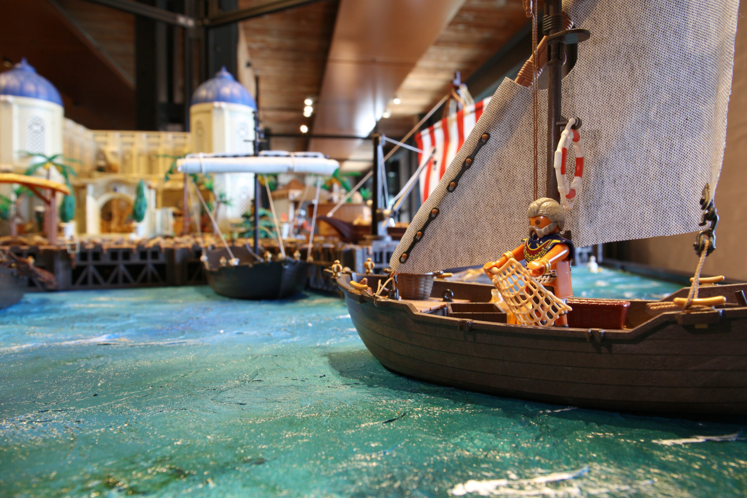 Spielzeugsegelboot mit Playmobil-Figur, Netz und Rettungsring auf blauer Wasserfläche vor Modellstadt im Hintergrund