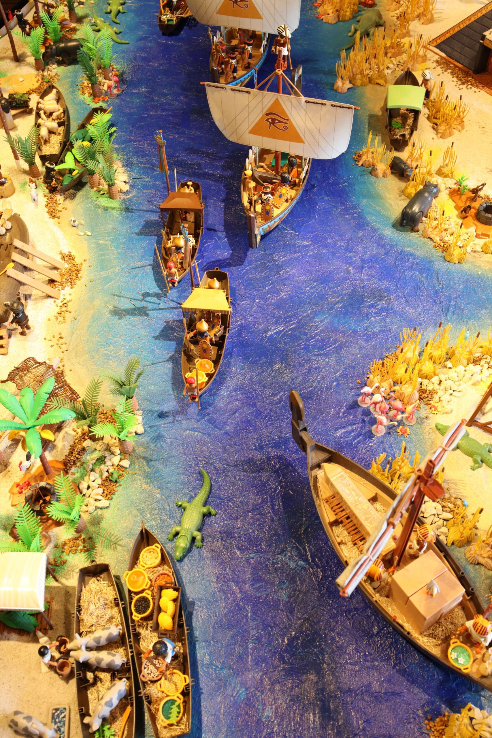 Bunte Spielzeugszenerie mit Segelschiff und Kanus auf blauem Fluss, Ufer mit Palmen, Tieren und Warenkähnen.