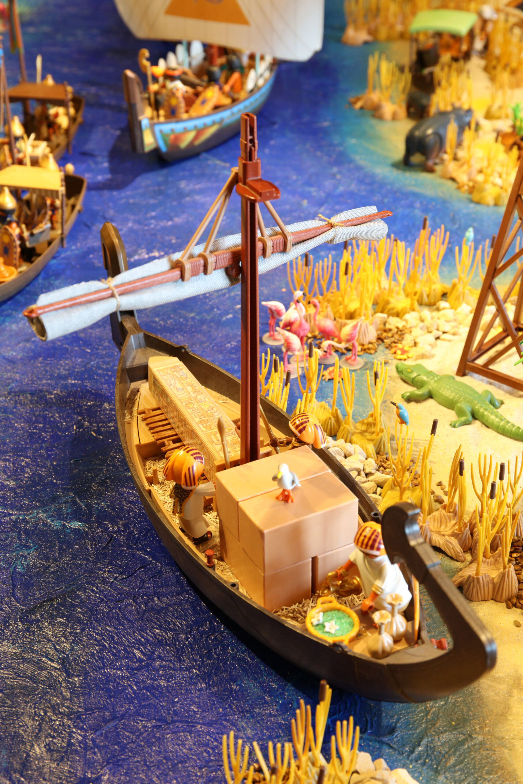 Spielzeug-Boot mit verpackter Ladung und Miniaturfiguren auf blauem Wasser vor gelbem Schilf und Tieren.