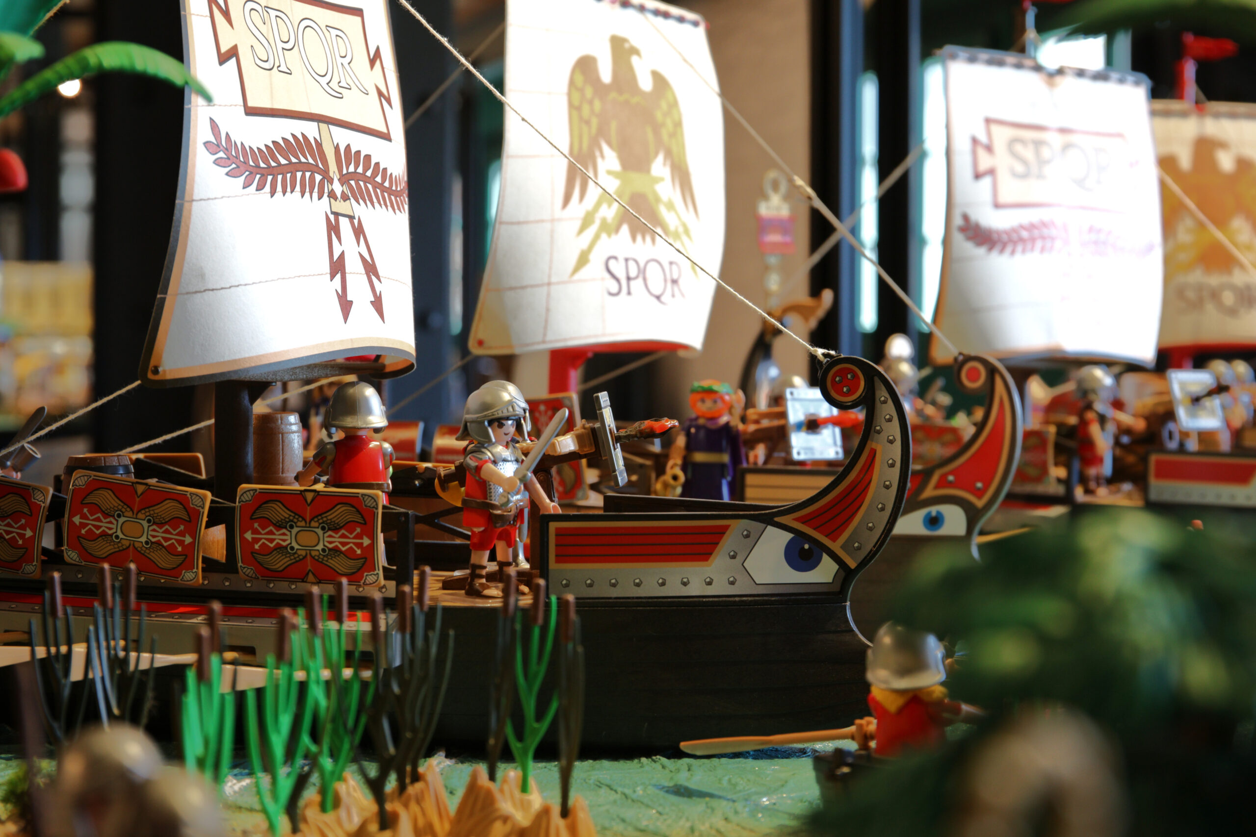Spielzeug-Römerschiffe mit SPQR-Segeln und Playmobil-Soldaten, vorn ein Schiffsruder mit Augenmalerei und gestaltete Schilde.