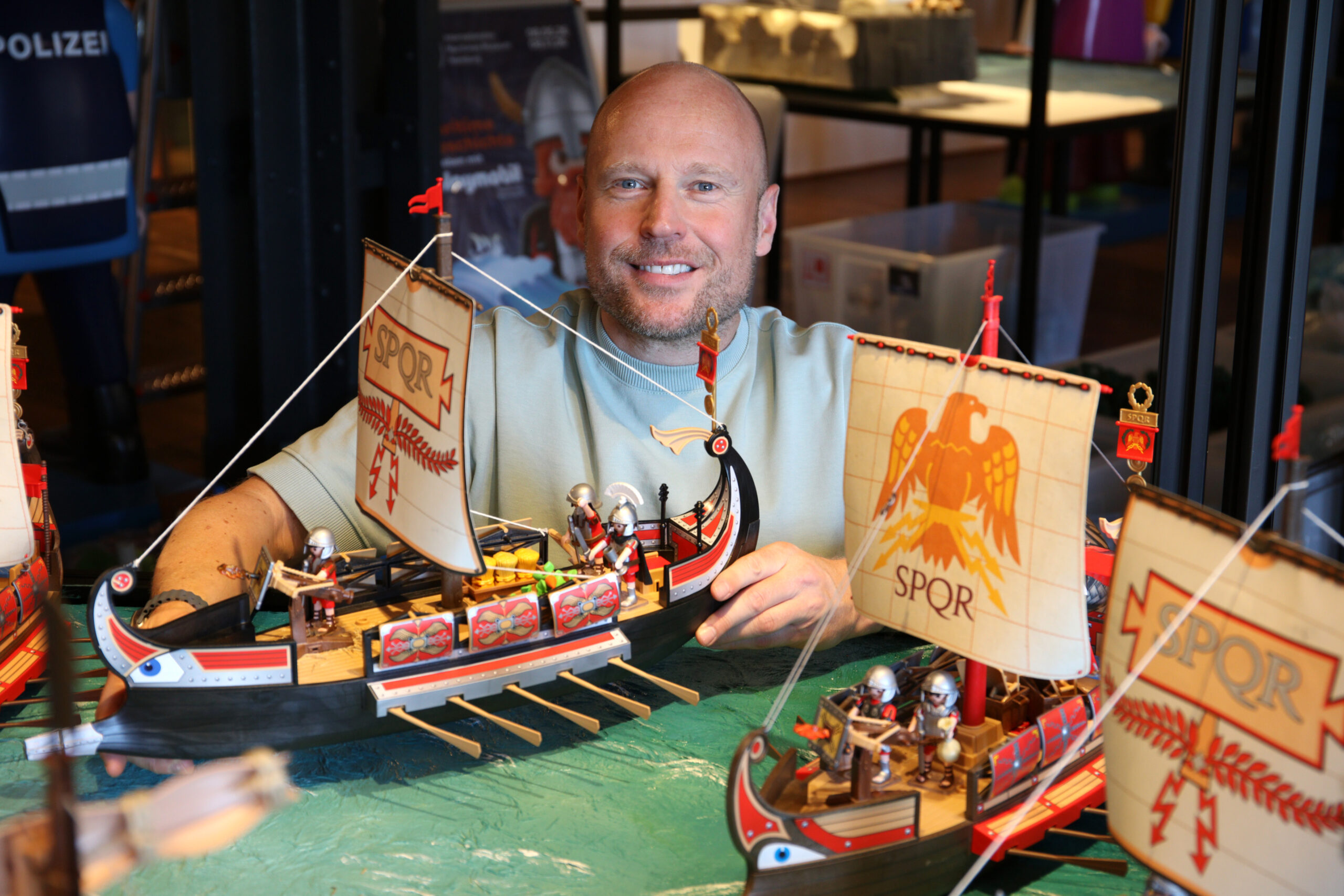Lächender Mann hinter detaillierten Spielzeug-Römerschiffen mit SPQR-Segeln und kleinen Römerfiguren auf grünem Untergrund.