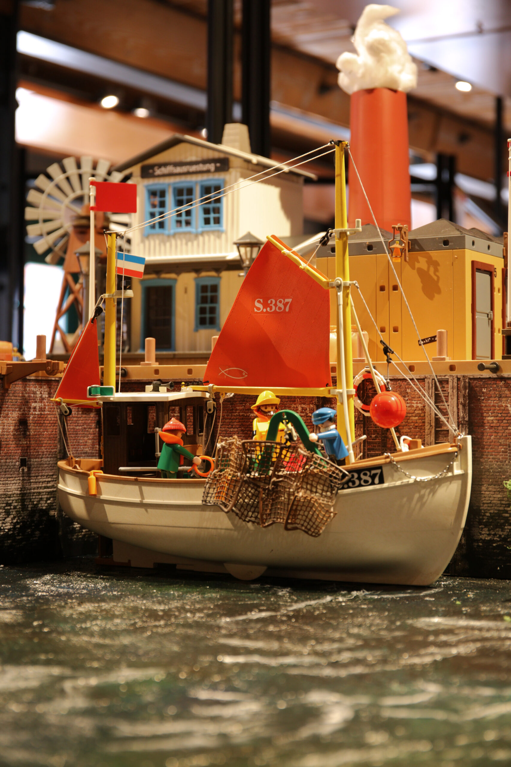Spielzeug-Fischerboot mit rotem Segel S.387, Mini-Figuren und Krabbenkörben im Hafen vor bunten Gebäuden.