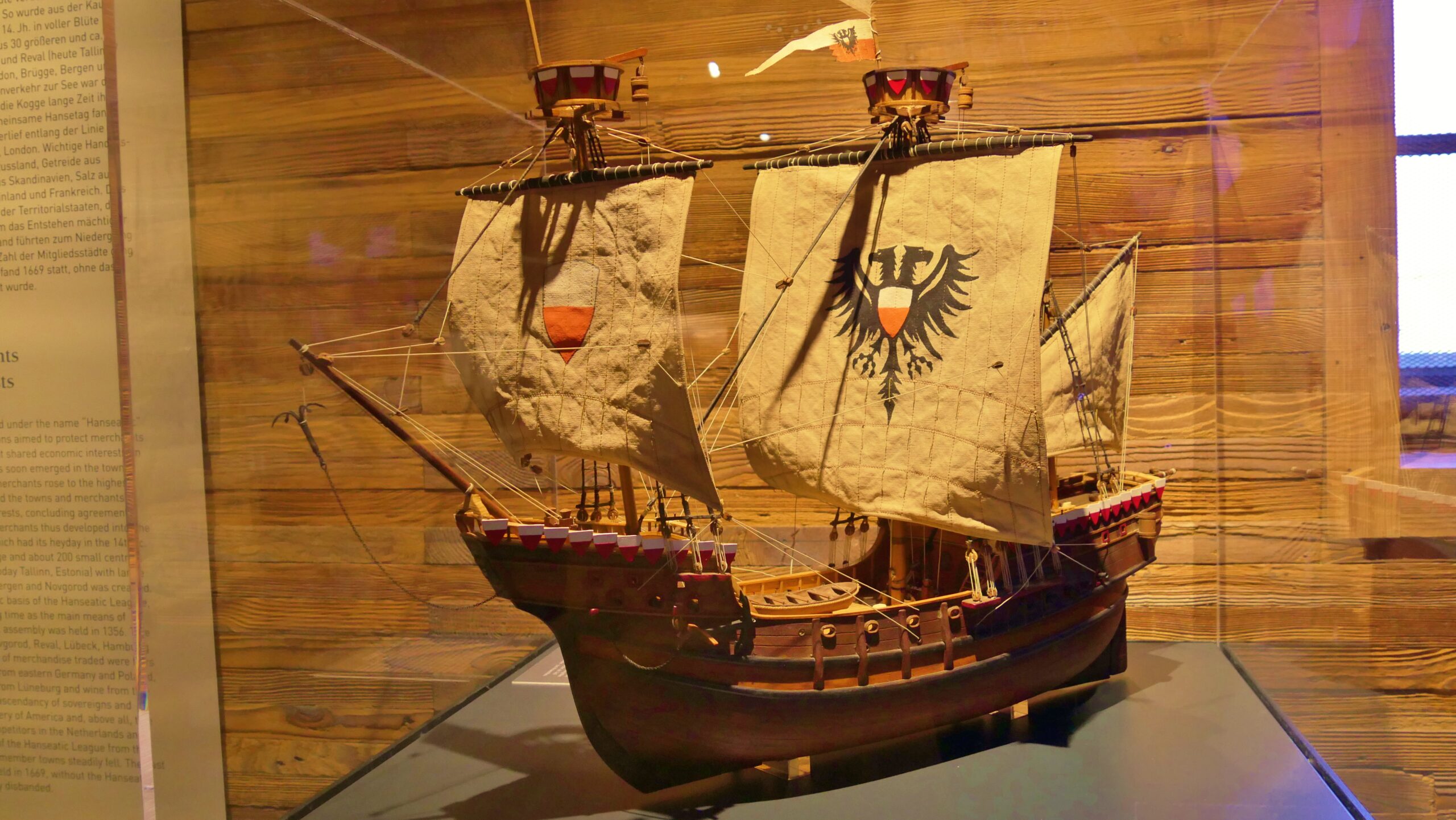 Zweimastiges Schiffsmodell mit leinenfarbenen Segeln, schwarzem Doppeladler und roten Schilden in einer Vitrine.