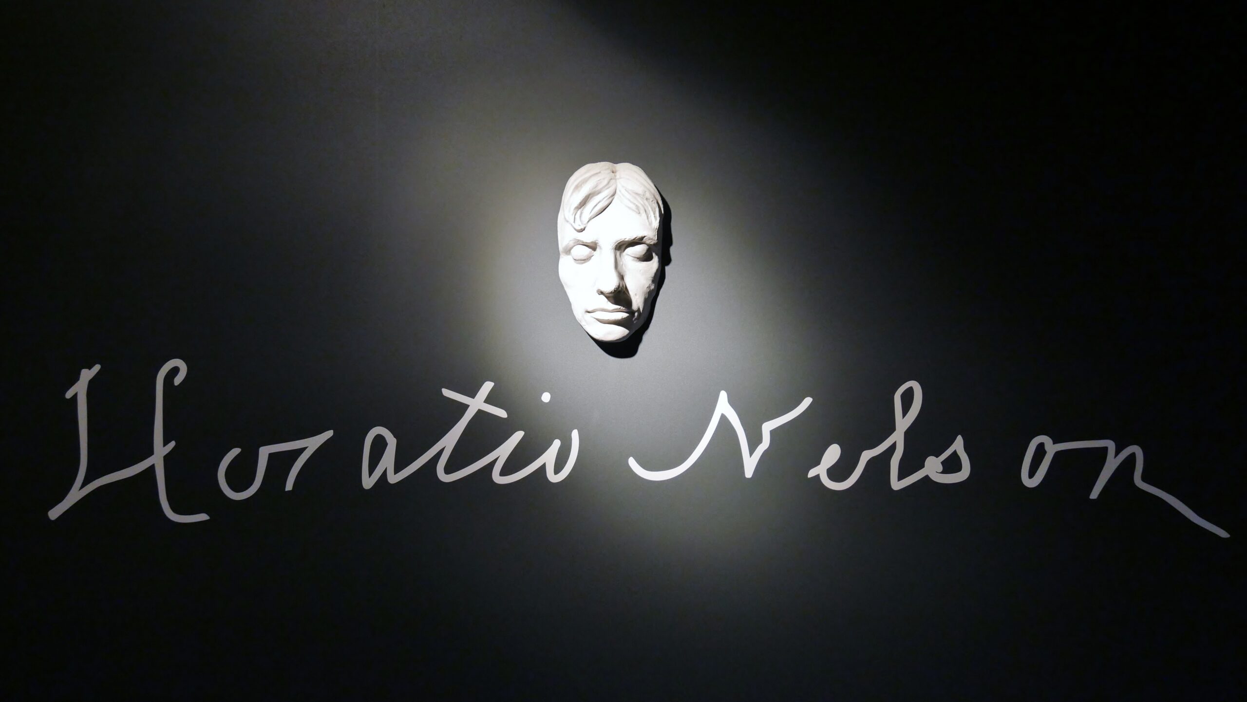 Weiße Gipsgesichtsmaske an dunkler Wand, von Scheinwerfer beleuchtet, mit handschriftlichem Schriftzug "Horatio Nelson" darunter.