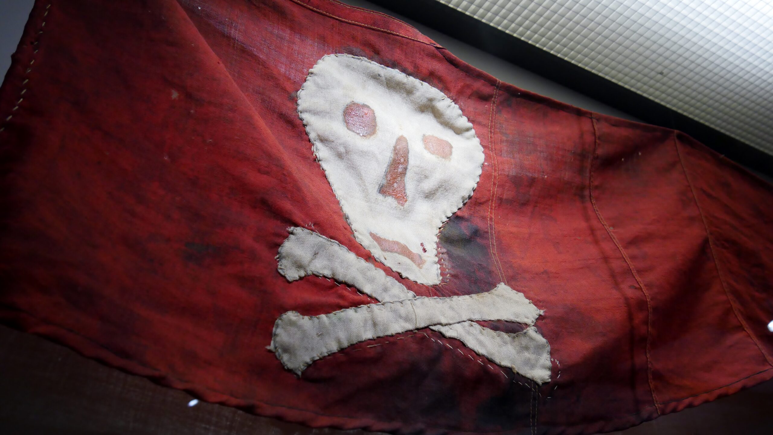 Rotes, abgenutztes Tuch mit aufgenähtem weißen Totenkopf und gekreuzten Knochen, ausgebleicht und fleckig.