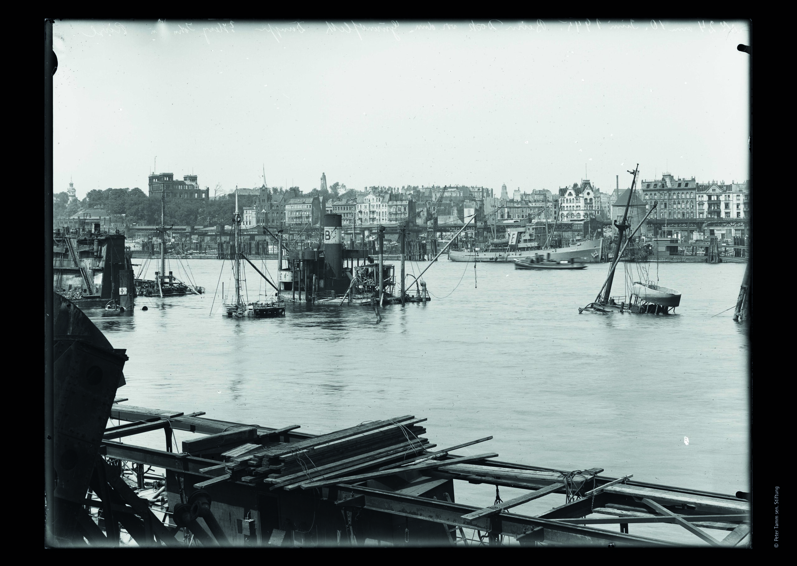 Hafenblick: Bauholz und Gerüst im Vordergrund, halb versunkene Boote und Arbeitsplattformen im Wasser, Stadtsilhouette im Hintergrund.