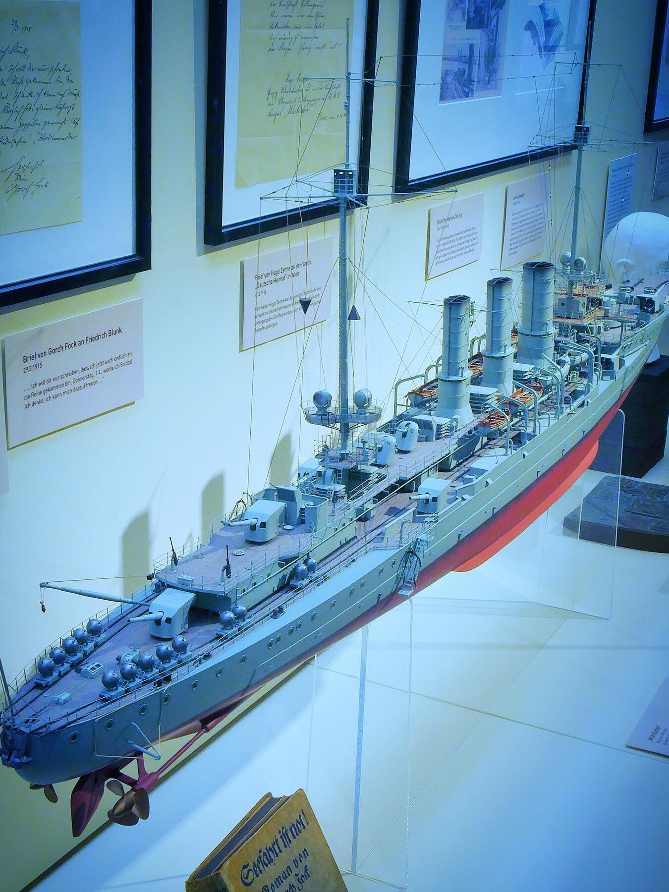 Detailliertes Modell eines Kriegsschiffs mit drei Schornsteinen, Masten und Takelage in Museumsvitrine vor gerahmten Dokumenten.