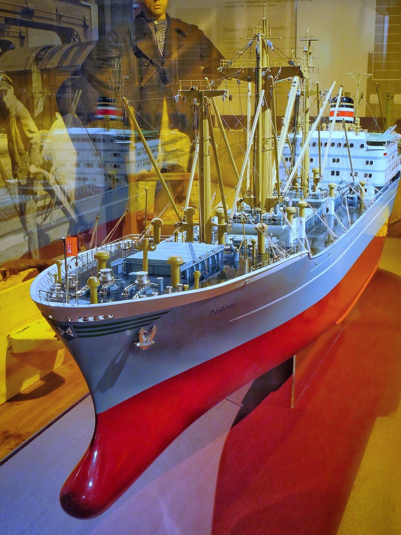 Detailliertes Modell eines Frachtschiffs mit rotem Rumpf, zahlreichen Ladebäumen, Takelage und Weißaufsatz, in Museumsvitrine mit Reflexionen.