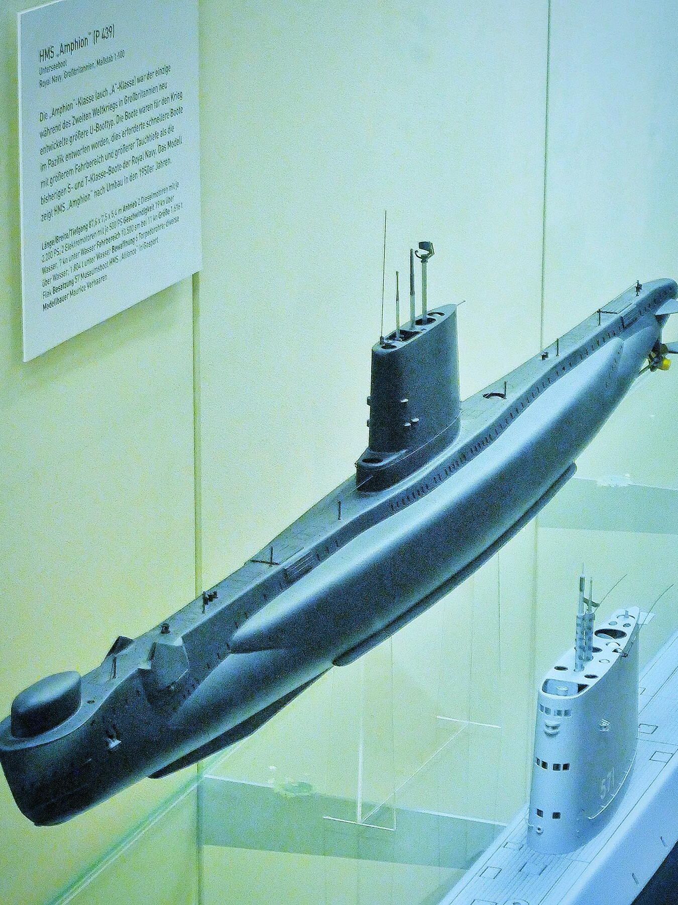 Modell eines grauen U-Boot-Rumpfes mit Turm, Antennen und Schraube in einer Vitrine, daneben Beschreibungszettel an der Wand