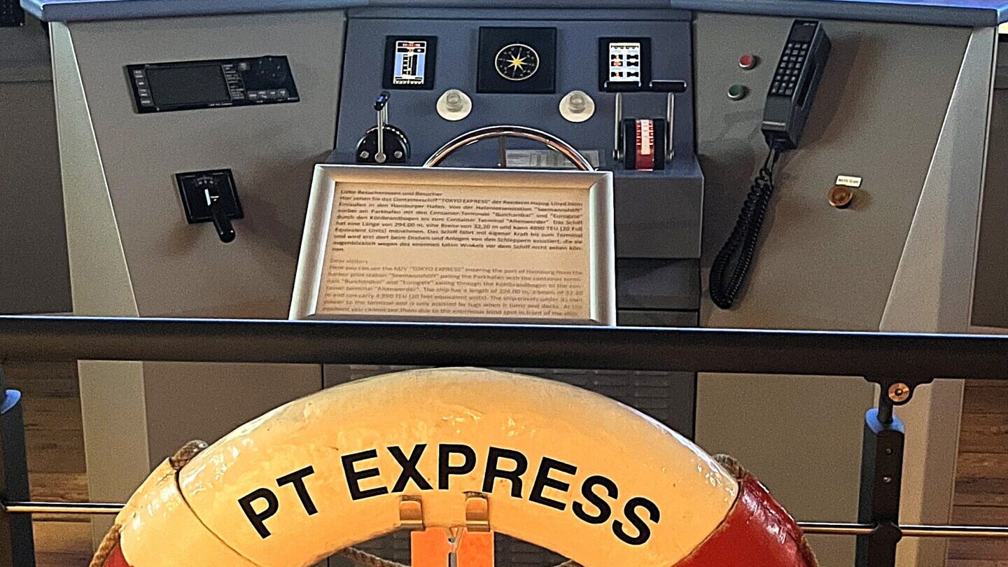 Rettungsring mit Aufschrift PT EXPRESS HAMBURG vor Steuerrad und mehreren Monitoren einer Schiffsbr&uuml;cke.