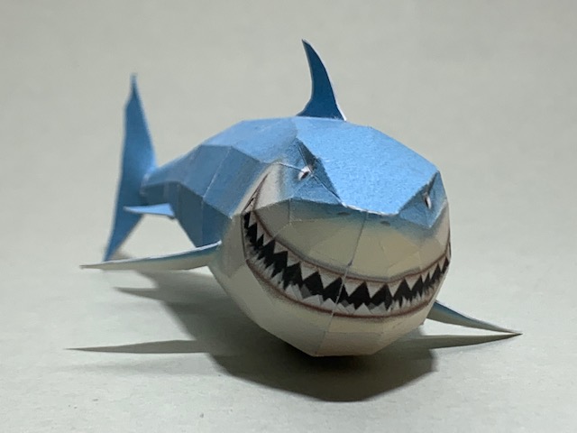 kleine gebastelte Papiershai mit gezacktem Maul, blauer Oberseite und wei&szlig;er Unterseite, frontal auf hellem Untergrund fotografiert