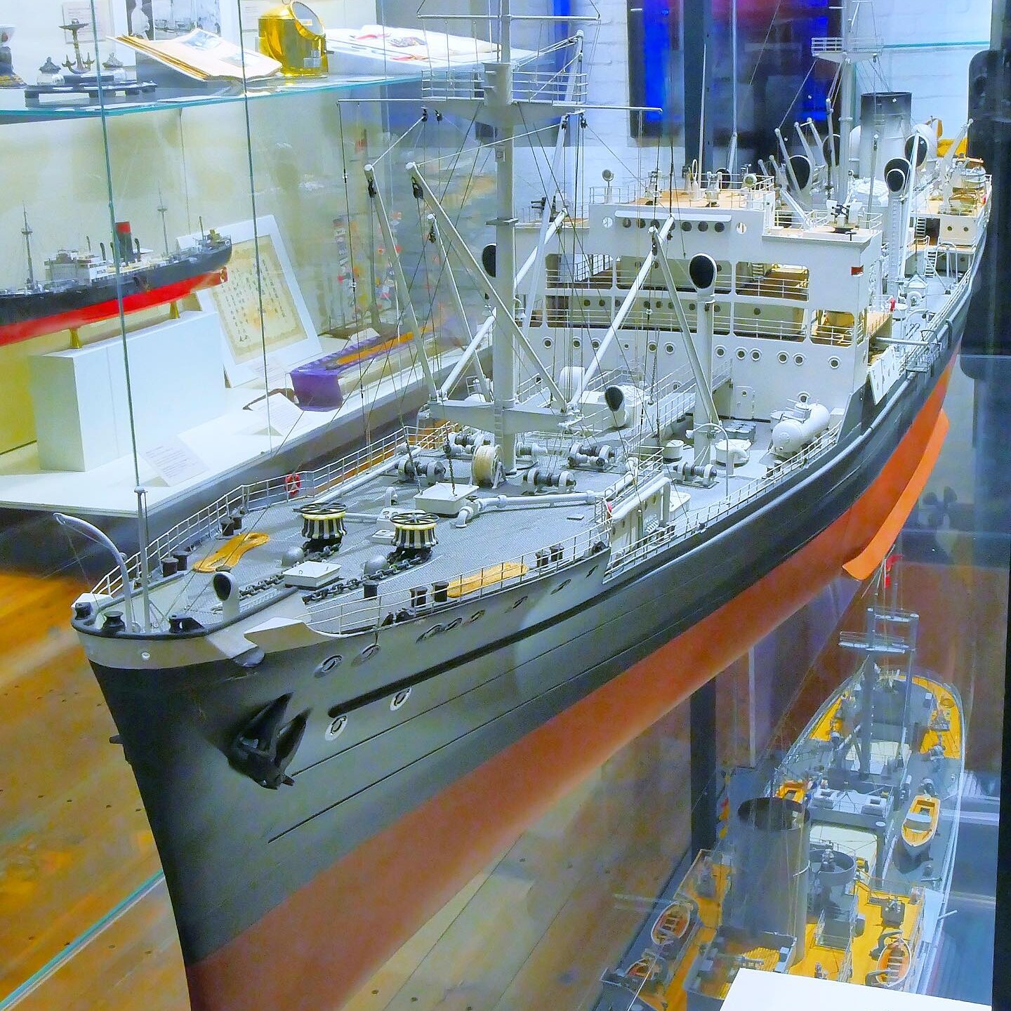 Detailliertes Schiffsmodell in Museumsvitrine mit Decksaufbauten, Kränen, Rettungsbooten und sichtbarem Rumpf in Rot und Schwarz.