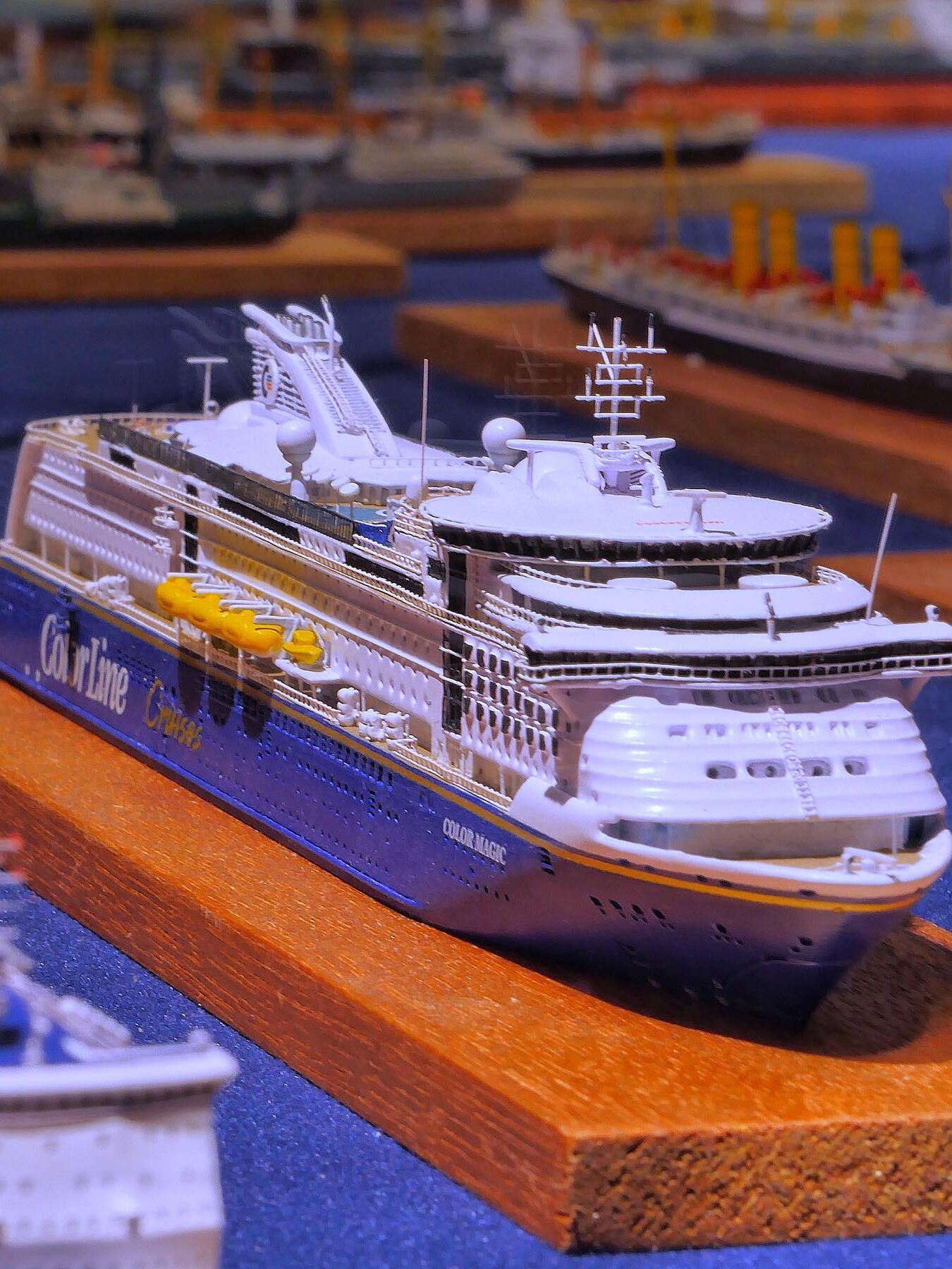 Weiß-blaues Modell eines großen Kreuzfahrtschiffs auf Holzsockel, Nahaufnahme mit Rettungsbooten und Details am Oberdeck.