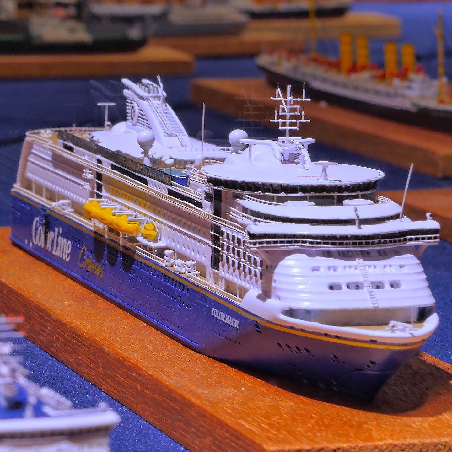 Weiß-blaues Modell eines großen Kreuzfahrtschiffs auf Holzsockel, Nahaufnahme mit Rettungsbooten und Details am Oberdeck.