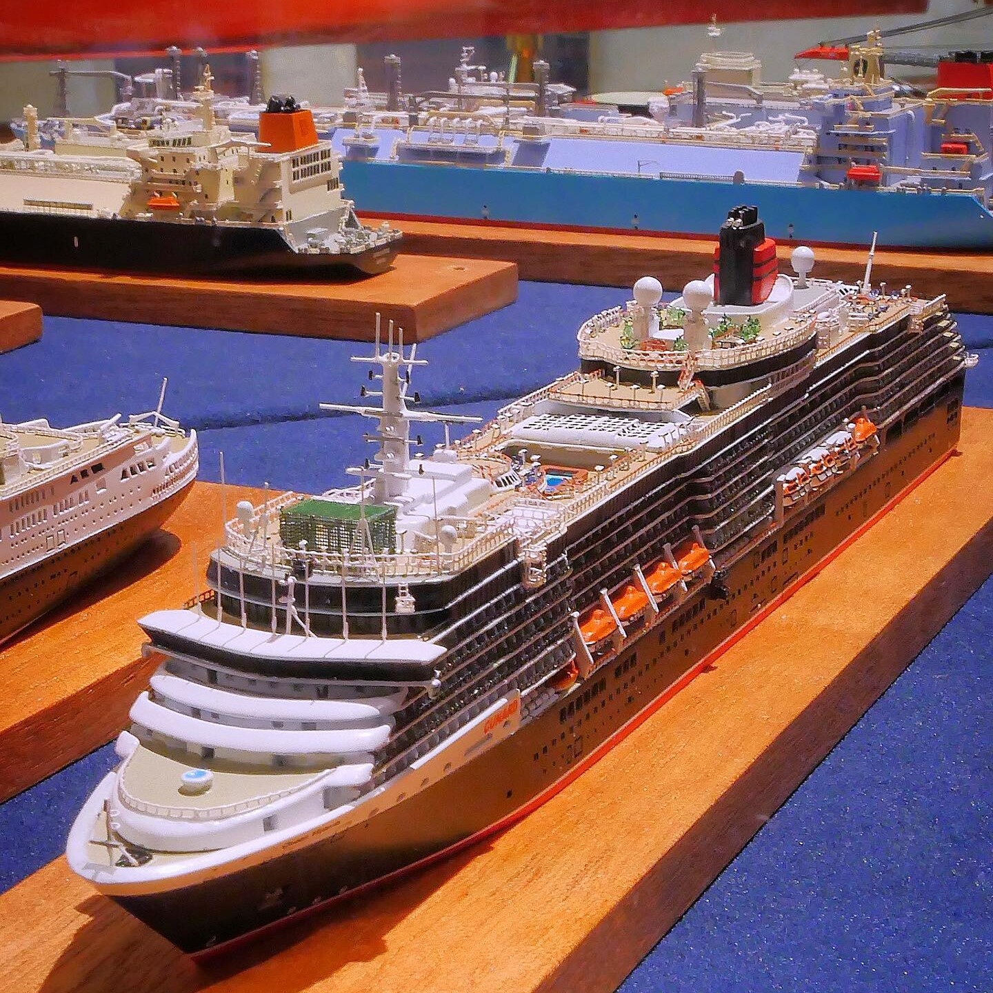 Detailliertes Modell eines großen Kreuzfahrtschiffs mit Deckpools, Rettungsbooten und Schornstein, ausgestellt auf Holzsockel neben weiteren Schiffsmodellen