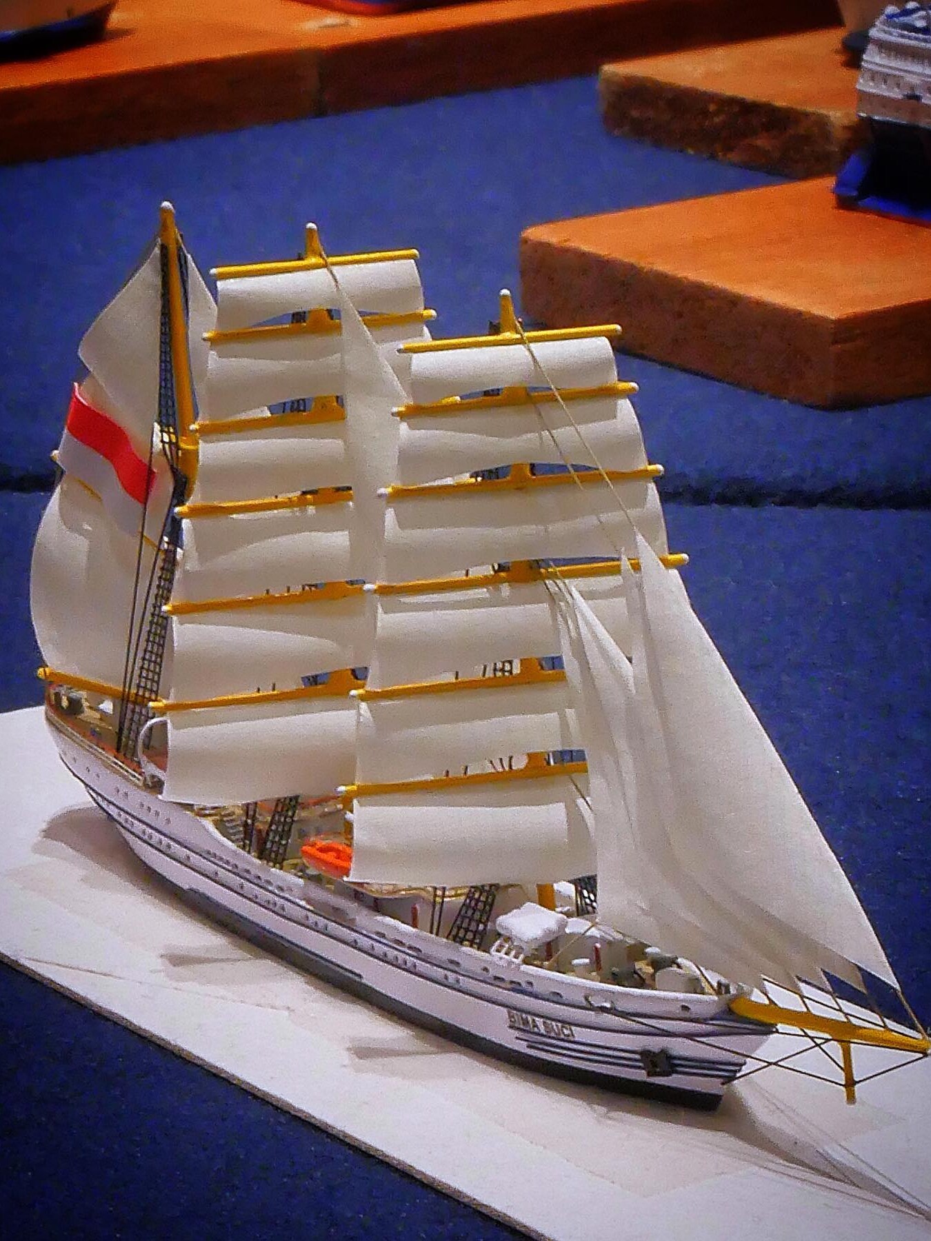 Weißes Modellsegelschiff mit mehreren eingerollten Segeln, gelben Rahen und roten Flaggenstreifen auf blauem Untergrund.