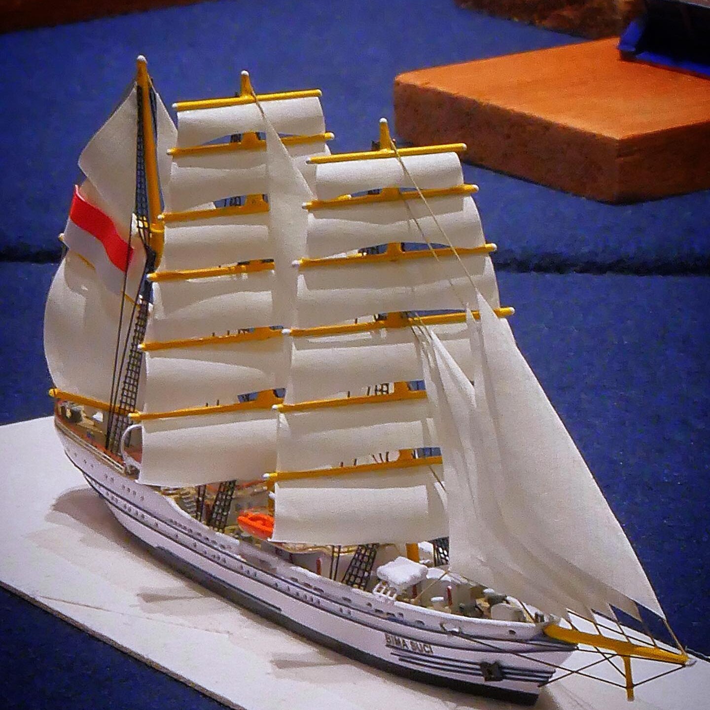 Weißes Modellsegelschiff mit mehreren eingerollten Segeln, gelben Rahen und roten Flaggenstreifen auf blauem Untergrund.