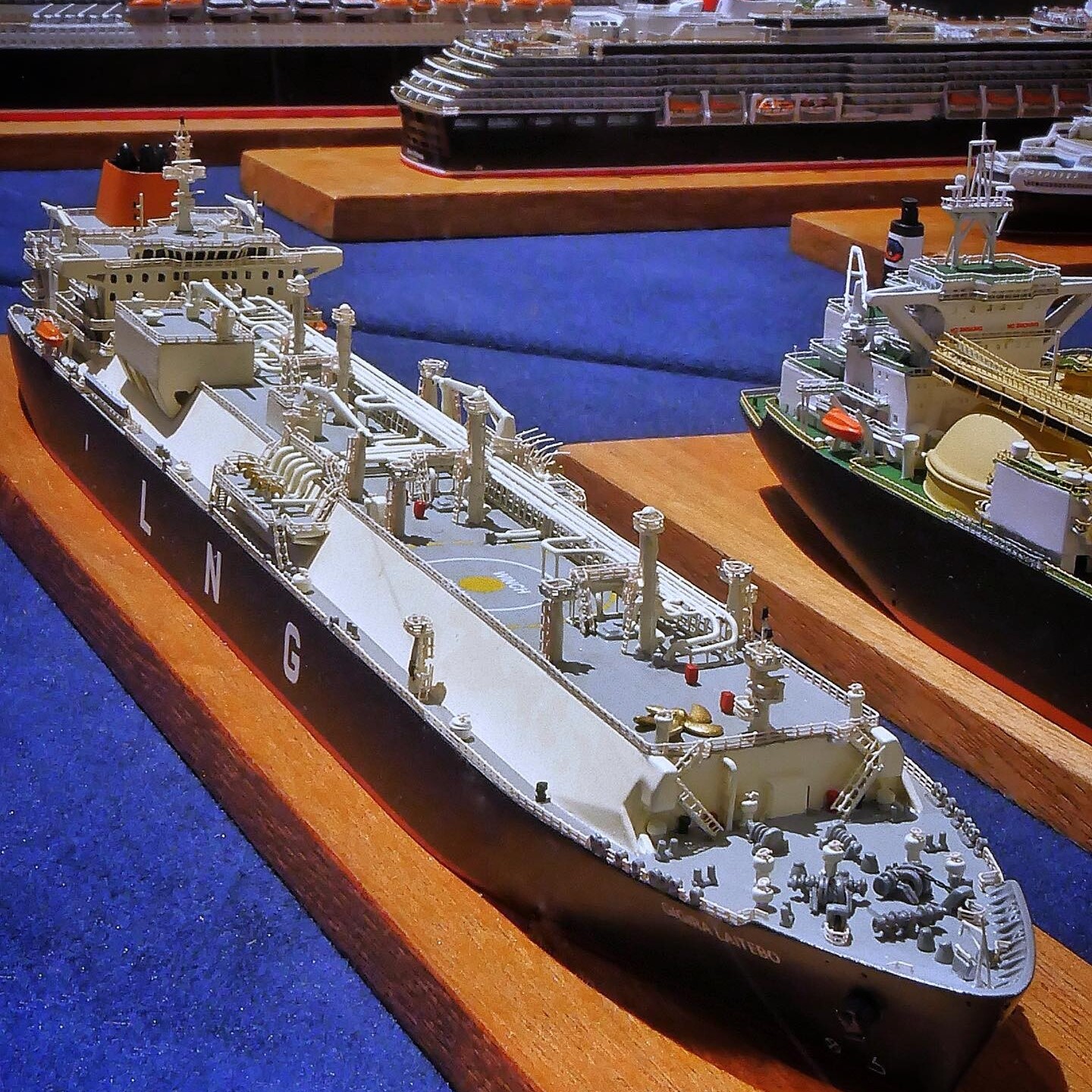 Mehrere detaillierte Schiffsmodelle auf Holzsockeln, vorn ein grauer Tankschiff-Prototyp mit Decksleitungen und Kräne, im Hintergrund Kreuzfahrtschiffe.